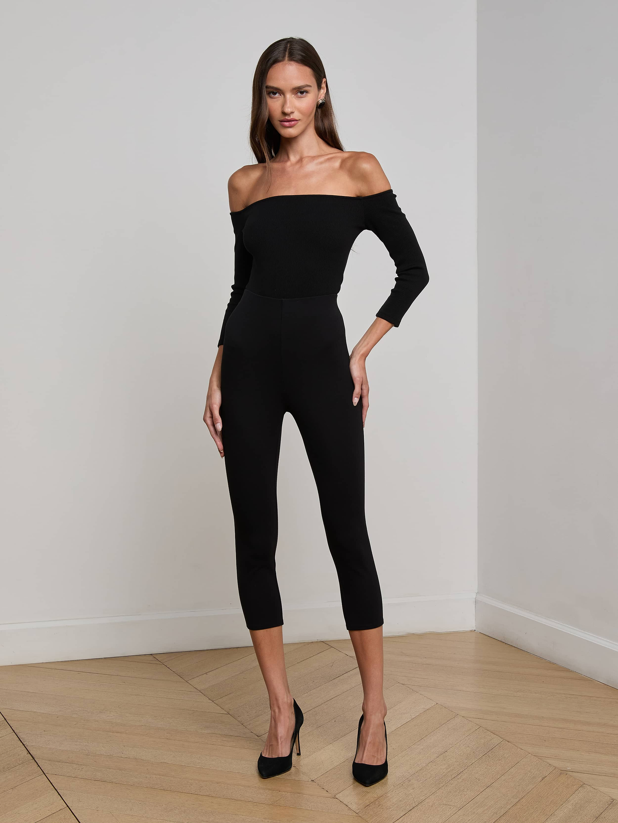 A woman in the Evelyn Off-The-Shoulder Bodysuit, featuring three-quarter sleeves and crafted from stretch cotton modal, pairs it with black heels as she stands against a plain white background, facing forward with one hand on her hip.