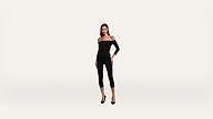 A woman in an off-the-shoulder black jumpsuit and black heels stands confidently against a plain white background.