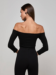 The woman, facing away from the camera, wears the Evelyn Off-The-Shoulder Bodysuit—crafted from stretch cotton modal—in black, paired with black pants as she stands against a plain light background.