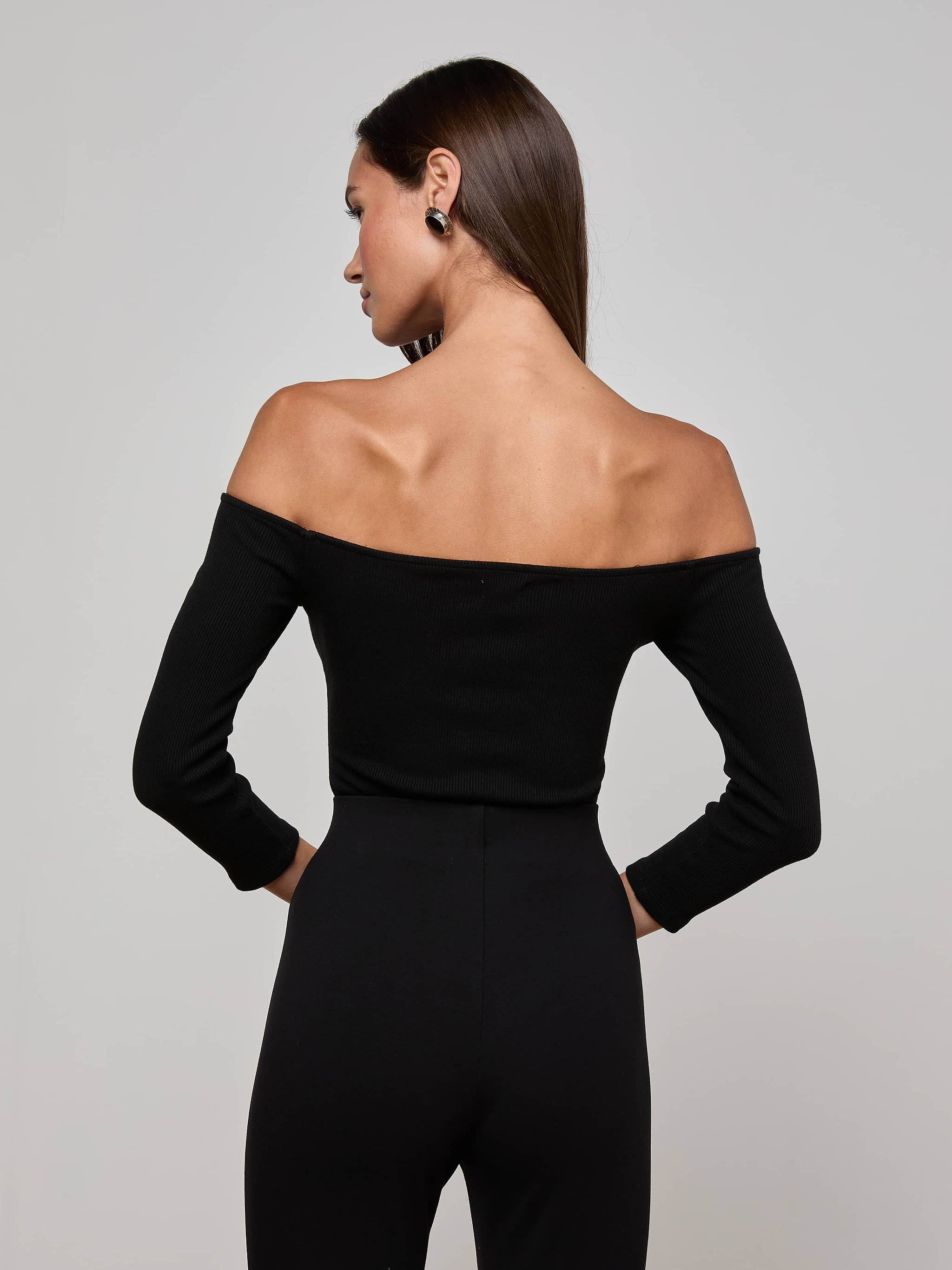 The woman, facing away from the camera, wears the Evelyn Off-The-Shoulder Bodysuit—crafted from stretch cotton modal—in black, paired with black pants as she stands against a plain light background.