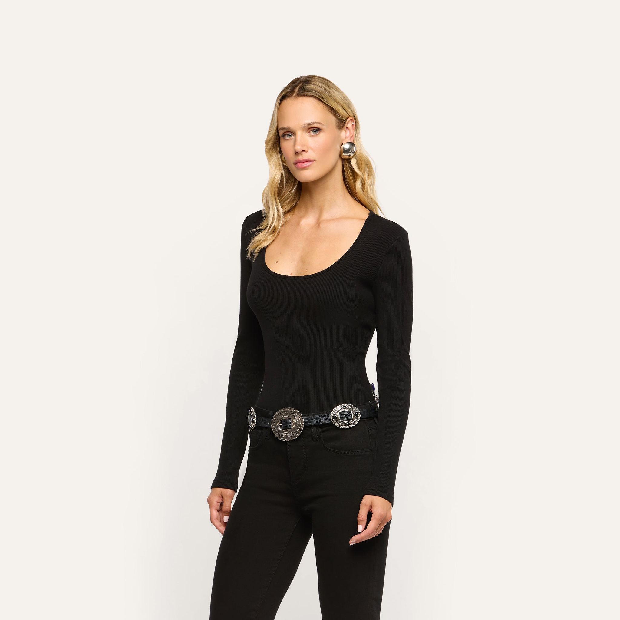 A woman wearing a long-sleeved black Elara Scoopneck Bodysuit in stretch cotton and black jeans with a statement silver concho belt.
