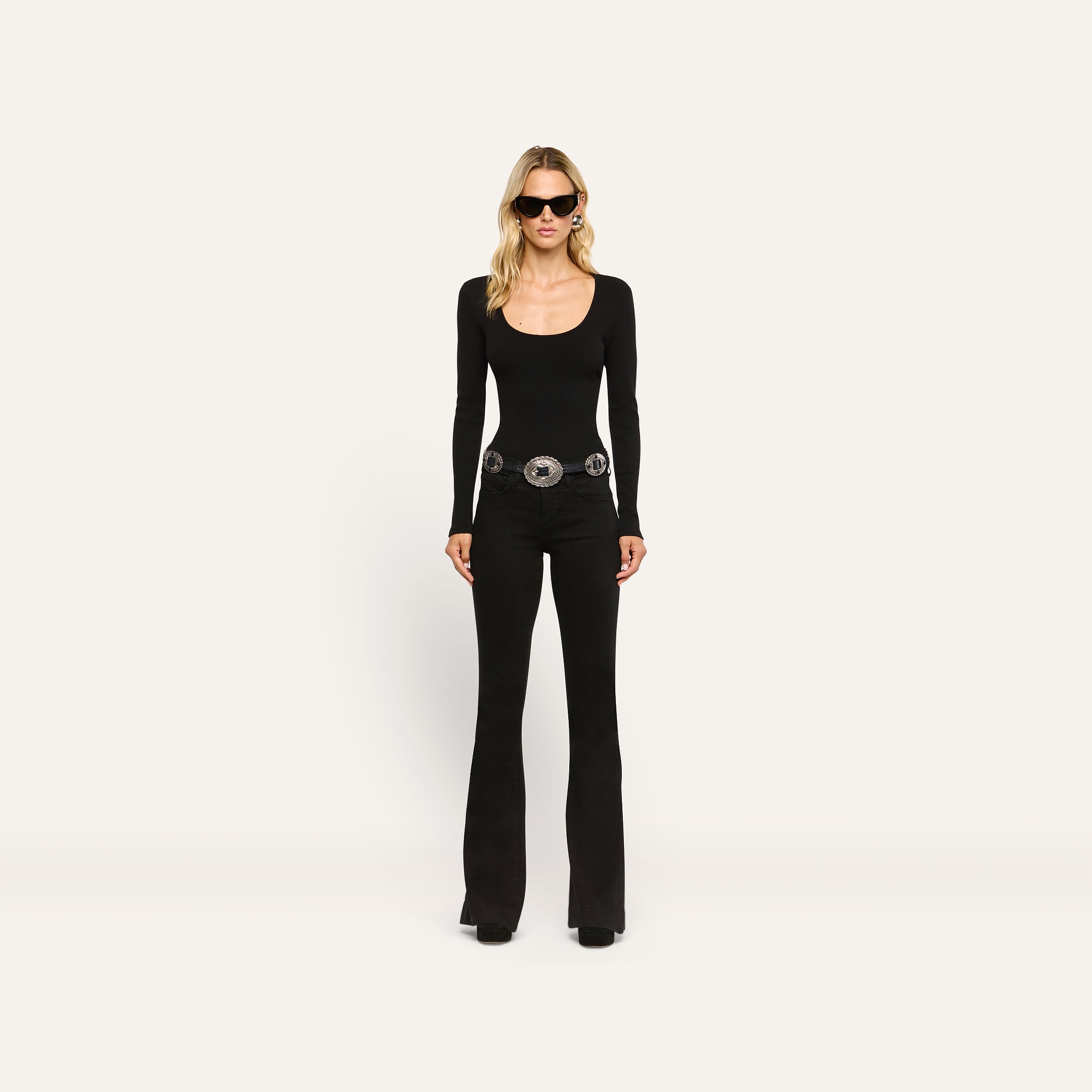 A woman wearing a black Elara Scoopneck Bodysuit, black flared jeans with a large silver statement belt, and dark sunglasses.