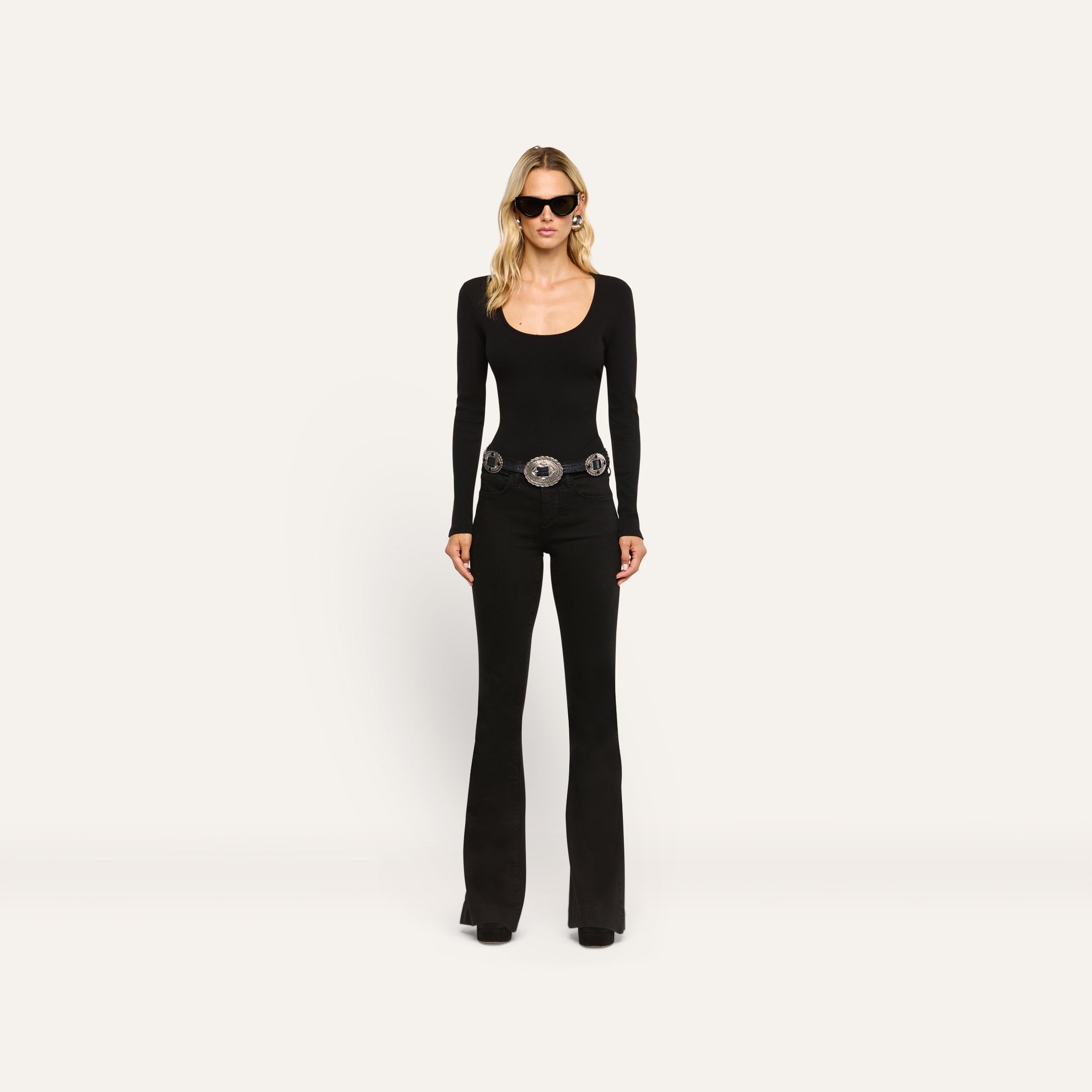 A woman wearing a black Elara Scoopneck Bodysuit, black flared jeans with a large silver statement belt, and dark sunglasses.