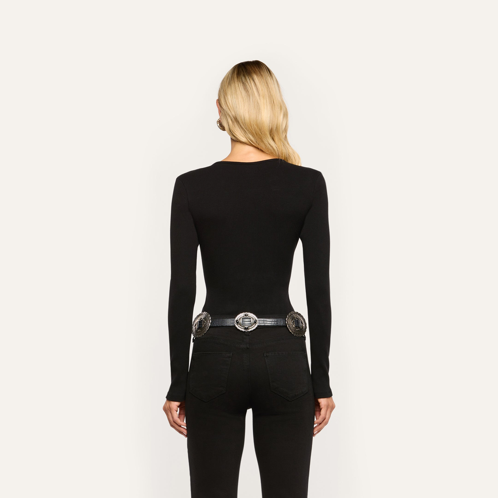 Back view of a person wearing a long-sleeved black Elara Scoopneck Bodysuit with black jeans and an ornate silver-buckle belt.