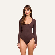A woman with long brown hair and bangs stands against a plain background, wearing the Elara Scoopneck Bodysuit—a long-sleeve, brown piece crafted from stretch cotton modal for a body-contouring fit. She looks directly at the camera with a neutral expression.