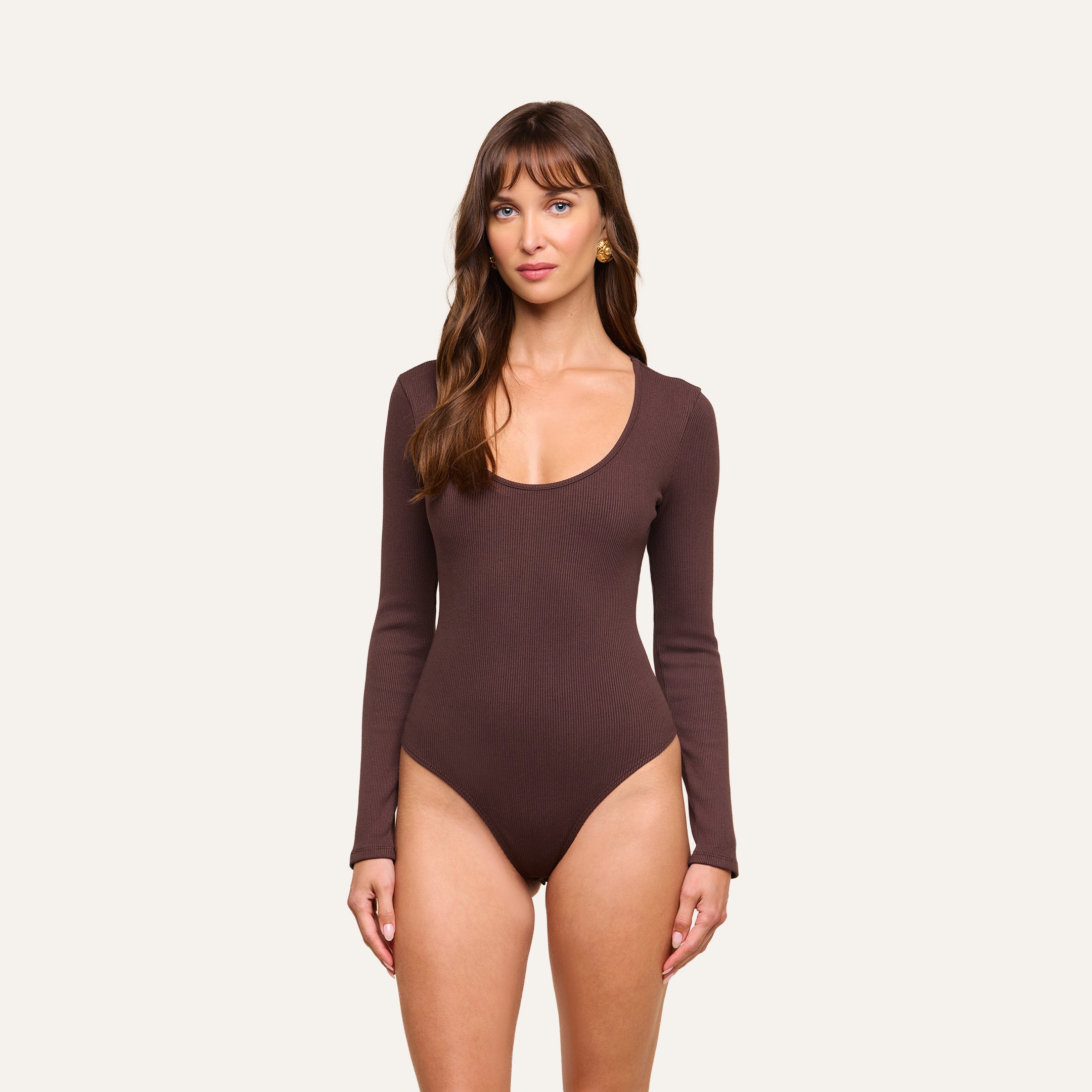 A woman with long brown hair and bangs stands against a plain background, wearing the Elara Scoopneck Bodysuit—a long-sleeve, brown piece crafted from stretch cotton modal for a body-contouring fit. She looks directly at the camera with a neutral expression.