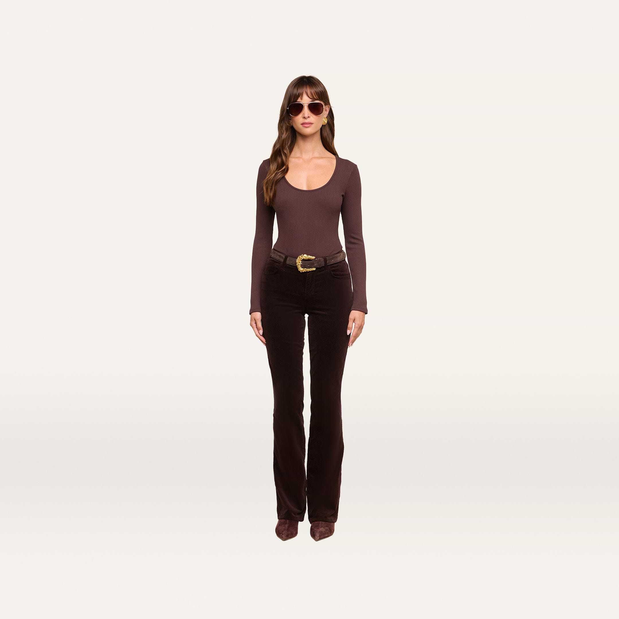 A woman stands against a plain white background, wearing sunglasses, the Elara Scoopneck Bodysuit—a long-sleeve brown stretch cotton modal piece that offers a body-contouring fit—paired with flared dark brown pants, a wide belt, and brown shoes.