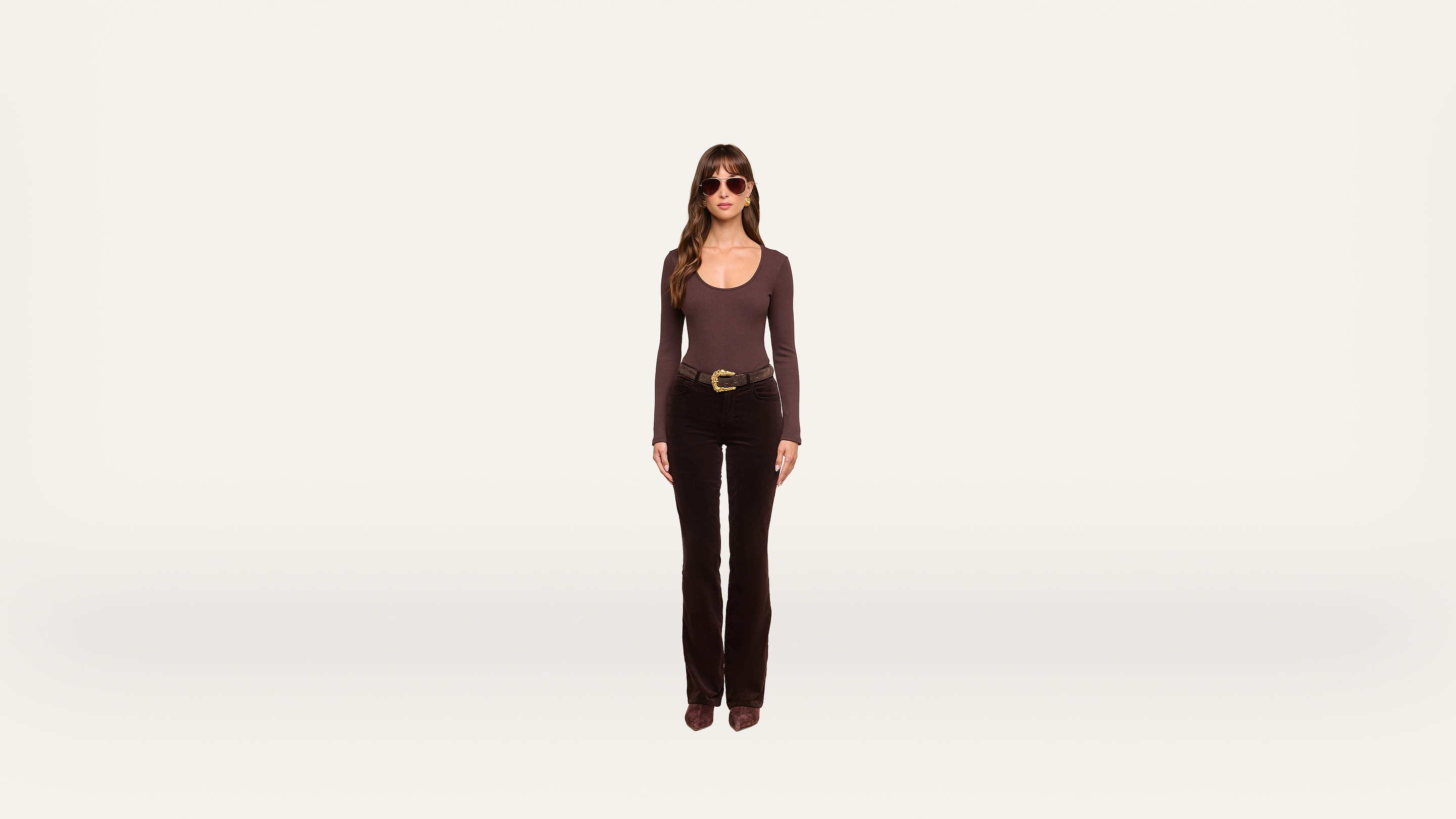 A woman wearing sunglasses, a long-sleeve top, black pants, and boots stands facing forward against a plain white background.