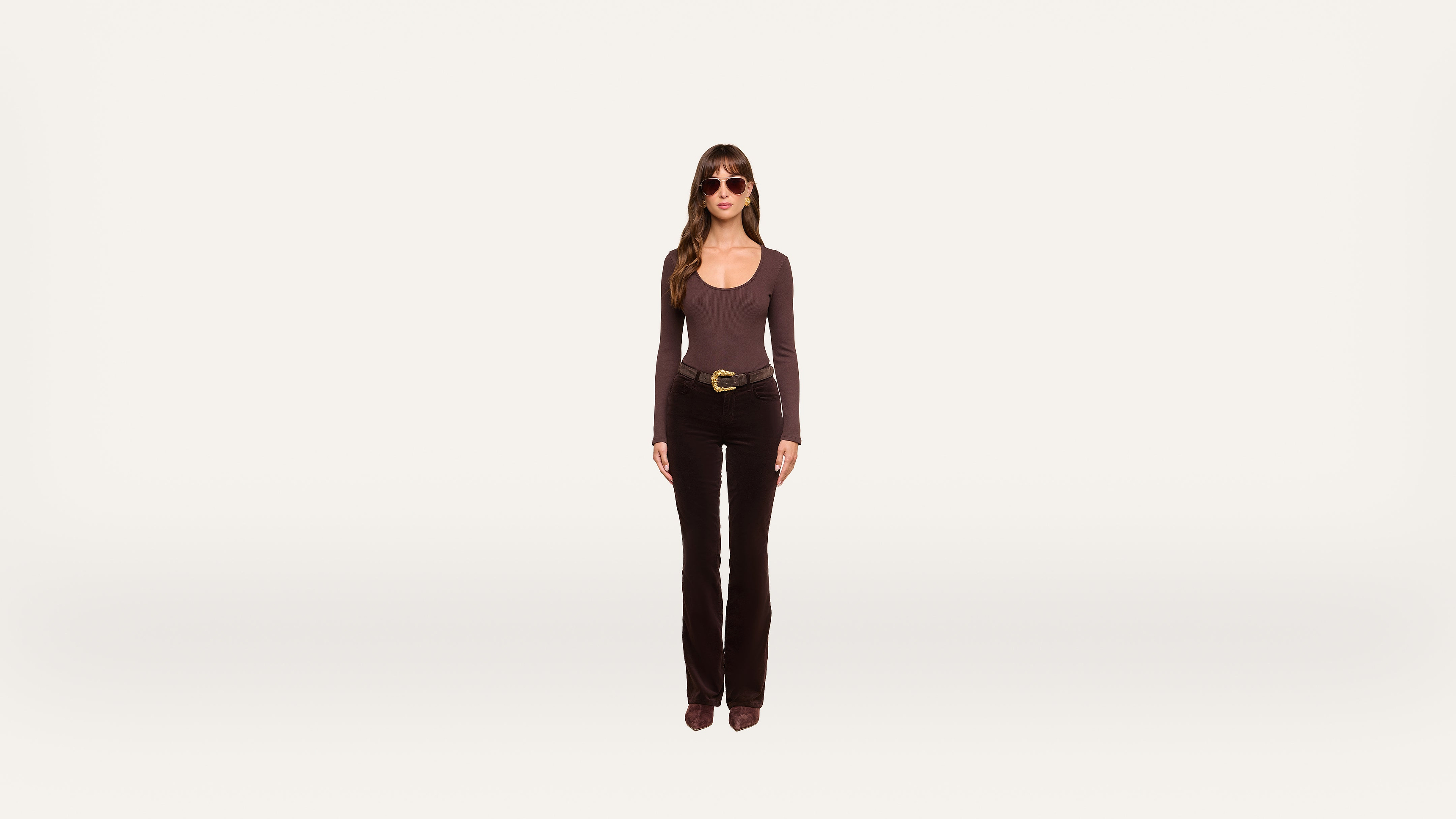 A woman wearing sunglasses, a long-sleeve top, black pants, and boots stands facing forward against a plain white background.