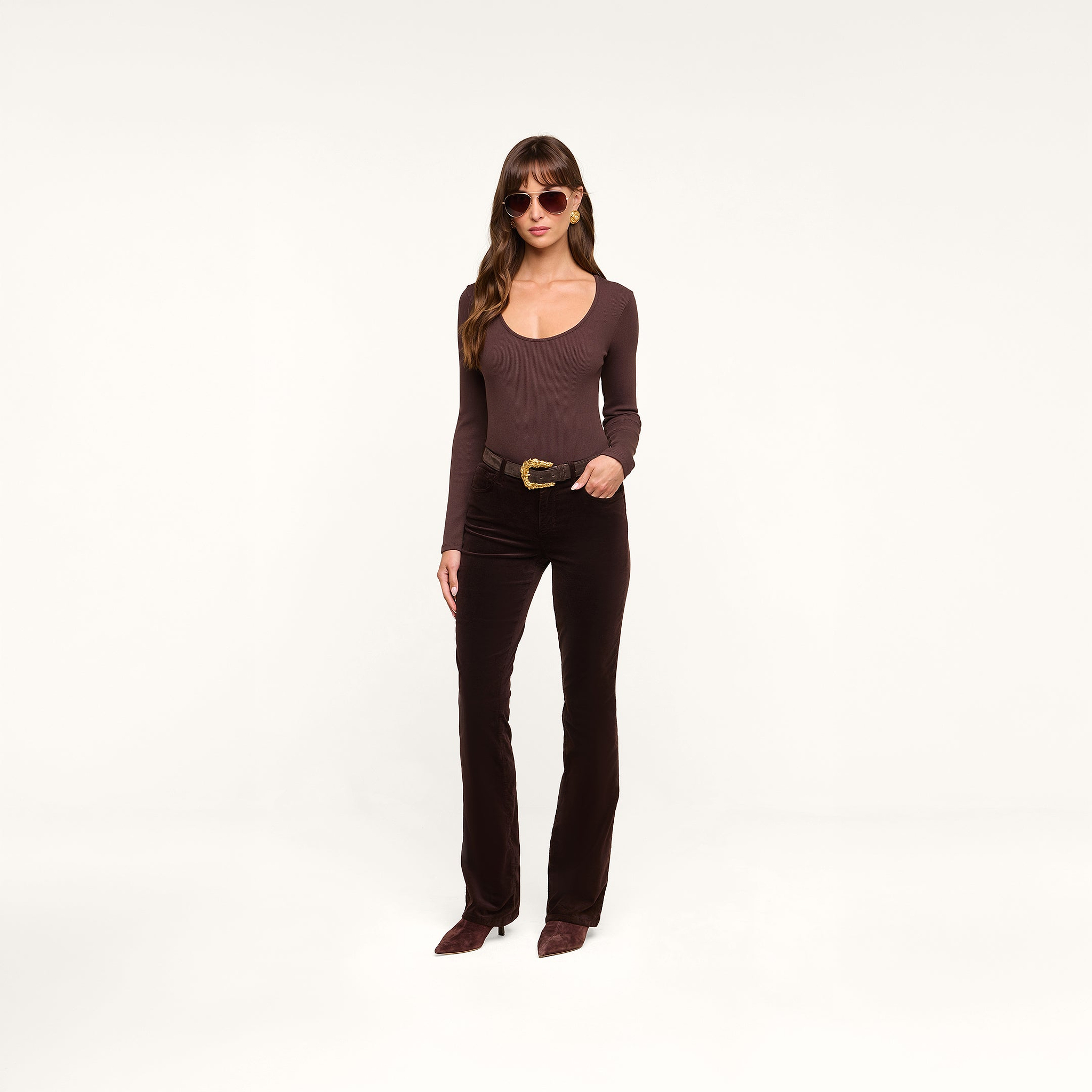 Wearing the Elara Scoopneck Bodysuit in a body-contouring brown hue, paired with matching brown pants and accessorized with brown sunglasses, a woman stands against a plain white background with one hand on her hip.