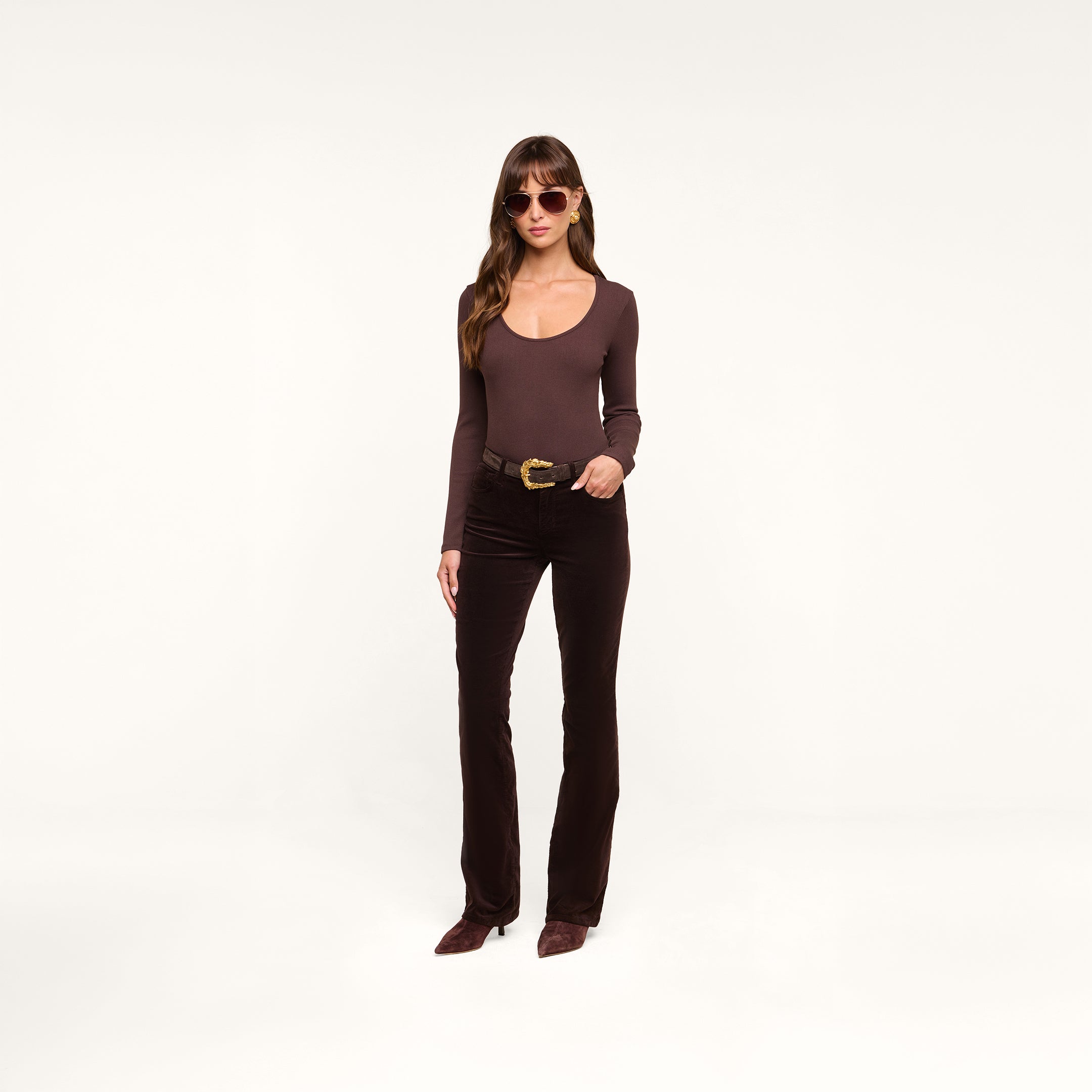 Wearing the Elara Scoopneck Bodysuit in a body-contouring brown hue, paired with matching brown pants and accessorized with brown sunglasses, a woman stands against a plain white background with one hand on her hip.