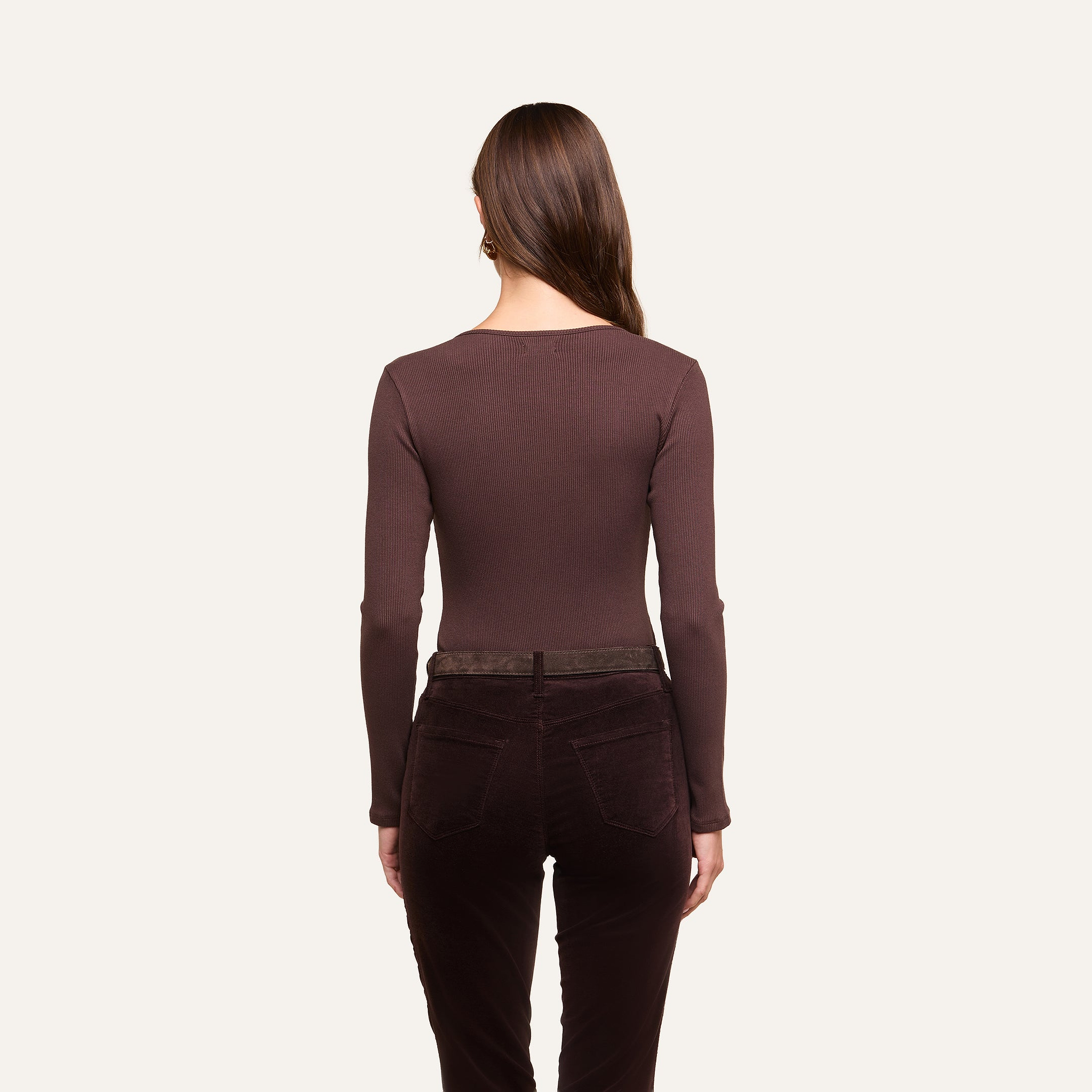 A woman with long brown hair, seen from behind, wears the Elara Scoopneck Bodysuit in a body-contouring fit along with dark brown pants against a plain off-white background.