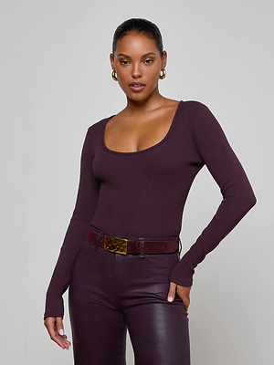 A woman stands confidently with one hand on her hip, wearing the L'AGENCE Elara Scoopneck Bodysuit in dark purple and matching high-waisted pants, styled with gold hoop earrings and a wide belt.