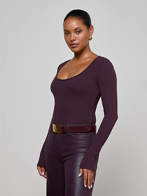 A woman wearing the L'AGENCE Elara Scoopneck Bodysuit—a fitted, long-sleeve, deep purple top—paired with matching high-waisted leather pants and a wide belt, stands against a plain light background.
