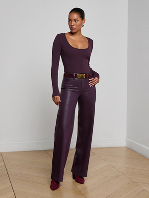 A woman stands confidently in a minimalist room with white walls and wooden floors, wearing the L'AGENCE Elara Scoopneck Bodysuit in stretch rib, styled with matching high-waisted wide-leg leather pants and heeled shoes.