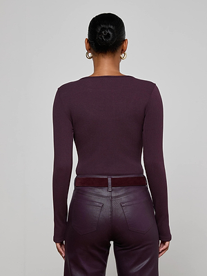 A woman with her hair in a bun and gold hoop earrings is shown from the back against a plain light gray background, wearing the L'AGENCE Elara Scoopneck Bodysuit paired with high-waisted dark burgundy leather pants.