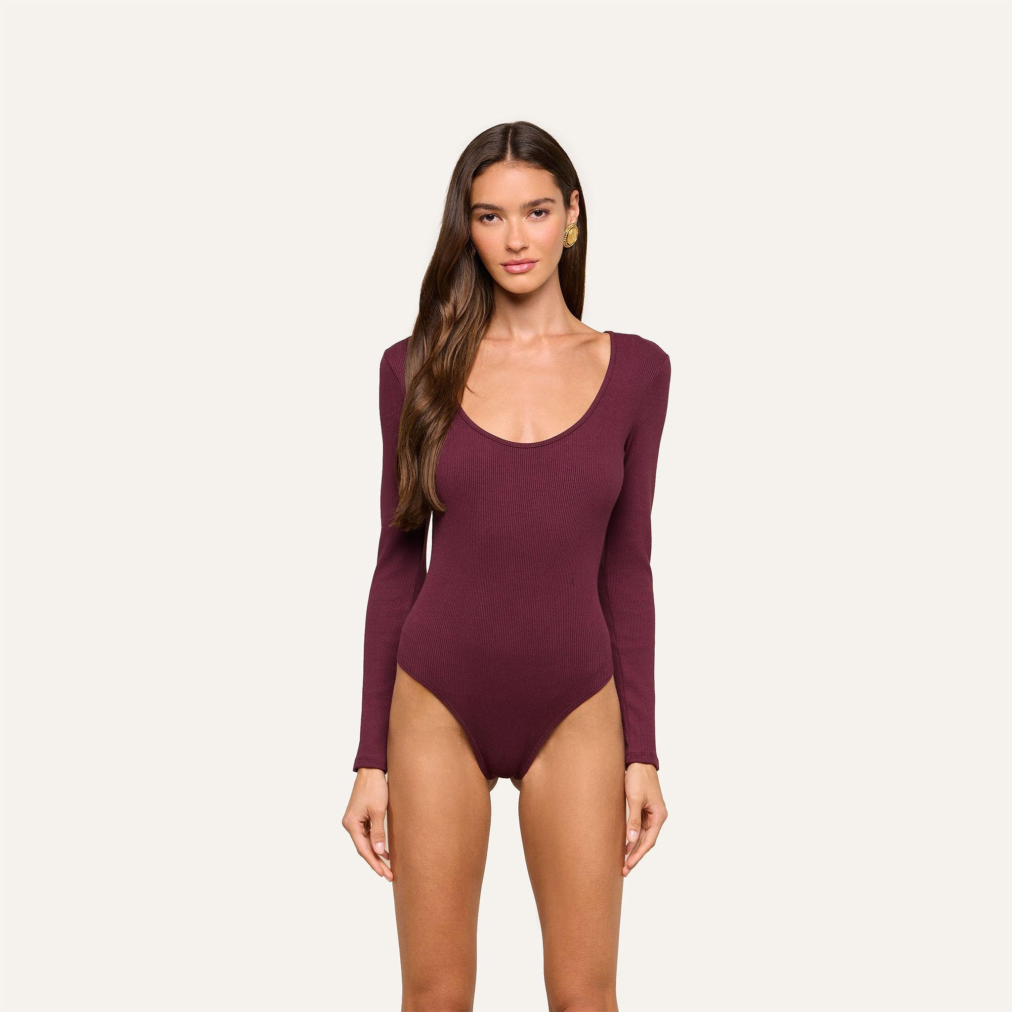A woman with long brown hair is wearing the Elara Scoopneck Bodysuit in deep burgundy, which features a body-contouring fit made from stretch cotton modal. She is standing against a plain, light background and looking straight at the camera with a neutral expression.