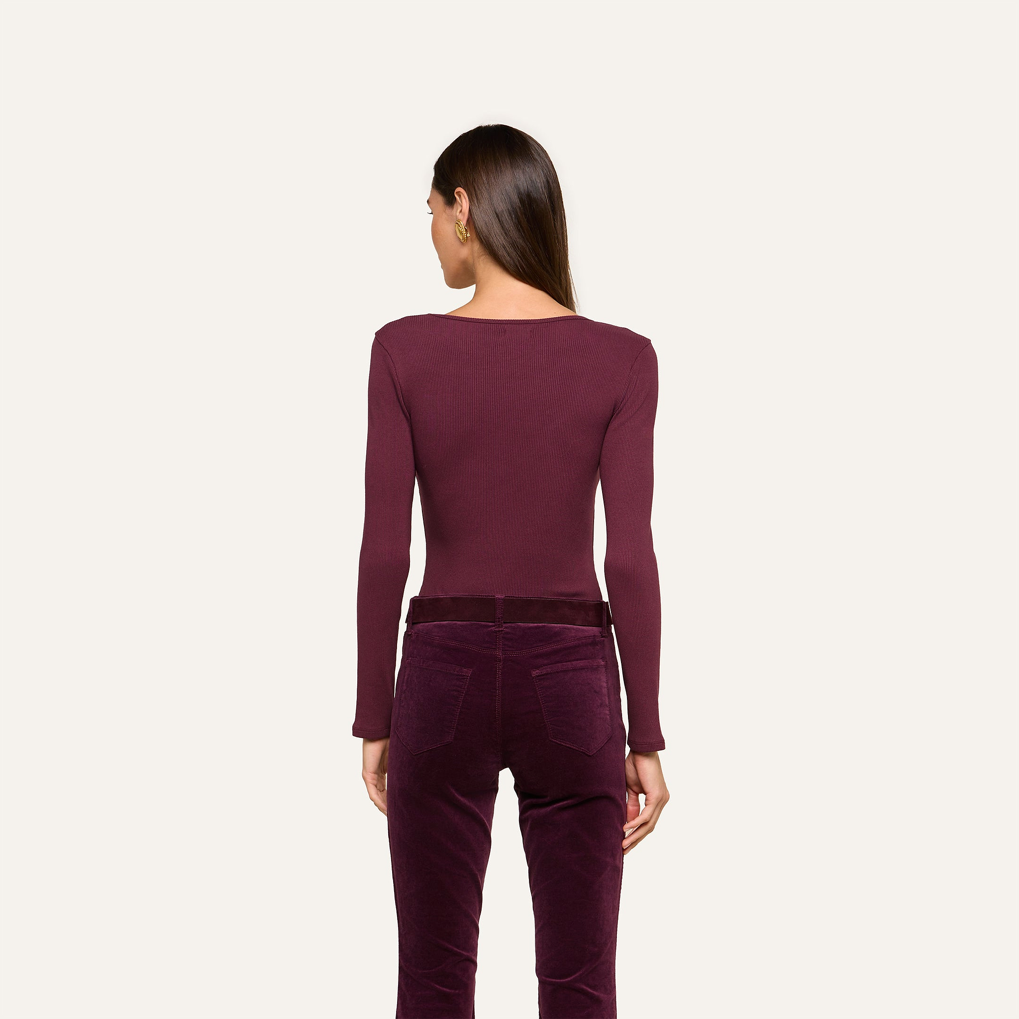 A woman stands with her back to the camera, wearing the Elara Scoopneck Bodysuit in maroon—crafted from stretch cotton modal—and high-waisted velvet pants in a coordinating shade, set against a simple light background. The look emphasizes the Elara’s sleek, body-contouring fit.