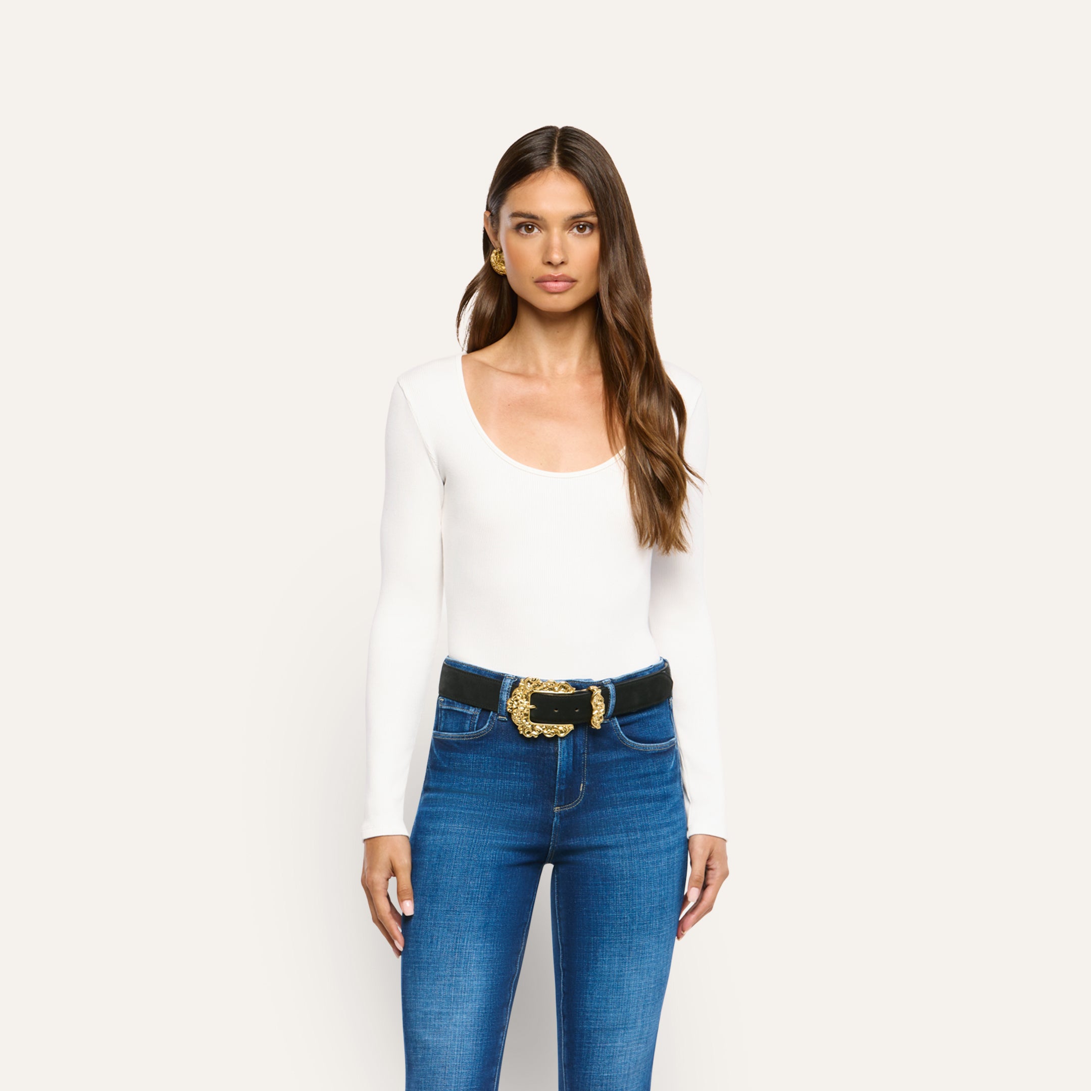 A woman wears an Elara Scoopneck Bodysuit in long-sleeved stretch cotton tucked into blue jeans with a black belt featuring an ornate gold buckle.