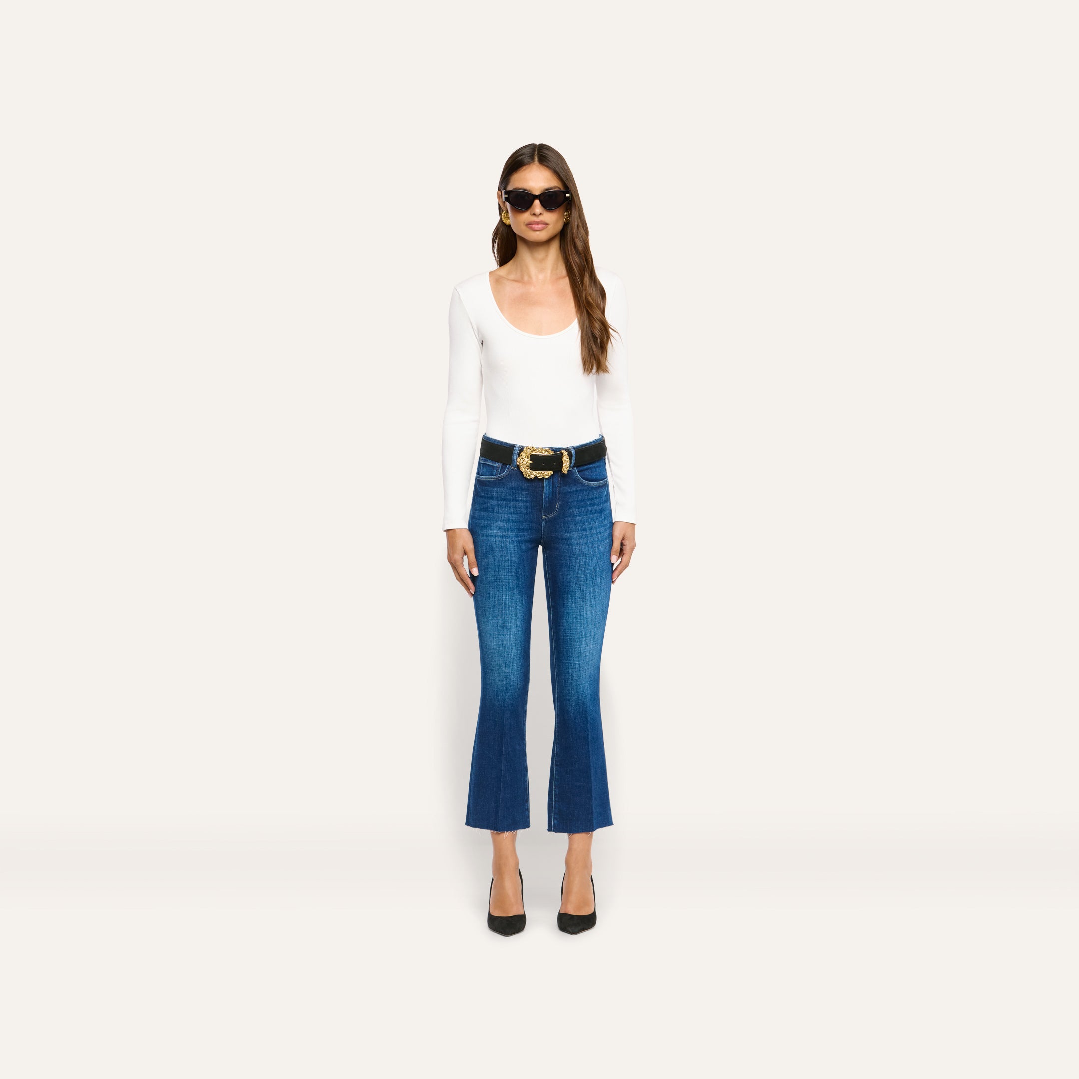 A woman wearing an Elara Scoopneck Bodysuit in white, dark blue cropped flare jeans, a black belt with an ornate gold buckle, and black heels.