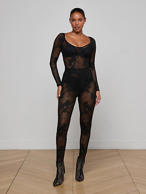 A woman poses against a white wall wearing the L'AGENCE Gwendolyn Mesh Knit Bodysuit with long sleeves and lace ankle boots on a wooden floor.