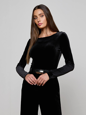 A woman with long brown hair models the L'AGENCE Seraphine Velvet Bodysuit, a black stretch velvet, long-sleeved top, paired with black pants. She stands against a plain light gray background with her hands clasped at her waist.