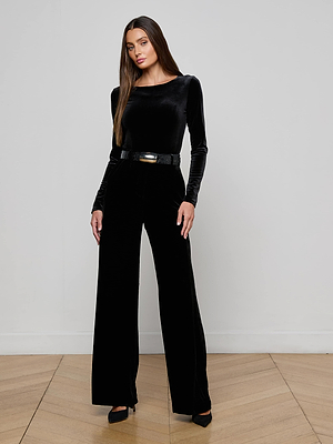 A woman with long, straight hair and a neutral expression stands confidently in a minimalist room, wearing the L'AGENCE Seraphine Velvet Bodysuit with wide-leg black pants, accessorized with a belt and black heels.