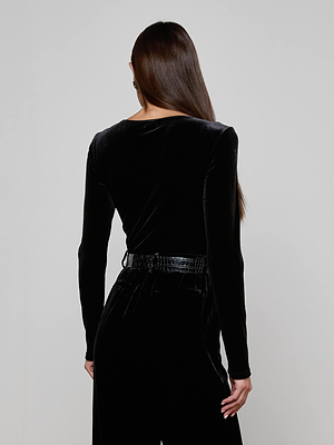 A woman with long brown hair faces away, wearing the Seraphine Velvet Bodysuit by L'AGENCE in black, paired with matching black pants against a plain light background.