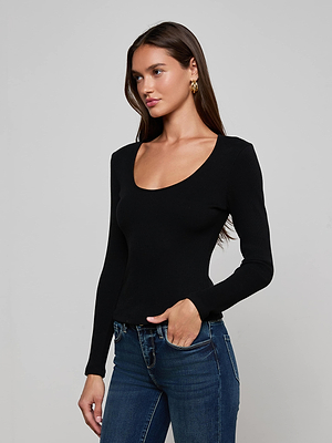 A woman with long brown hair wears the L'AGENCE Scarlet Scoopneck Tee— a fitted black long-sleeve top— and blue jeans. She stands against a plain light gray background, facing slightly to the side with a neutral expression.