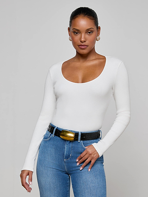A woman stands facing forward with one hand on her hip, wearing the L'AGENCE Scarlet Scoopneck Tee and blue jeans. Her hair is pulled back, she accessorizes with gold earrings, and poses against a plain light background.