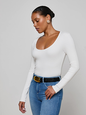 A woman in the L'AGENCE Scarlet Scoopneck Tee stands against a plain light background, wearing blue high-waisted jeans, a black belt with a gold buckle, and gold earrings, looking down with one hand on her hip.