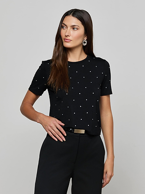 A woman with long brown hair wears the L'AGENCE Stormy Embellished Tee, styled with black trousers, a gold-buckled black belt, and statement earrings, standing against a plain light background.