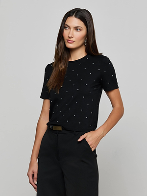 A woman with long brown hair stands against a plain background, wearing the L'AGENCE Stormy Embellished Tee and black high-waisted pants. One hand is in her pocket, and she gazes slightly to the side.