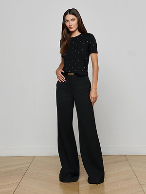 A woman with long brown hair stands indoors against a plain light wall, wearing the L'AGENCE Stormy Embellished Tee—a black cropped stretch rayon jersey top with small white polka dots—and high-waisted, wide-leg black pants.