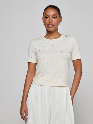 A woman stands against a plain light gray background wearing the Stormy Embellished Tee, a vintage-inspired white short-sleeve top with small dotted details, paired with a white pleated skirt.