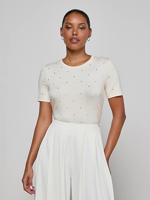 A woman stands against a plain background wearing the Stormy Embellished Tee, a vintage-inspired white fitted top with small black polka dots, paired with a high-waisted pleated white skirt and hoop earrings. She has a calm expression.