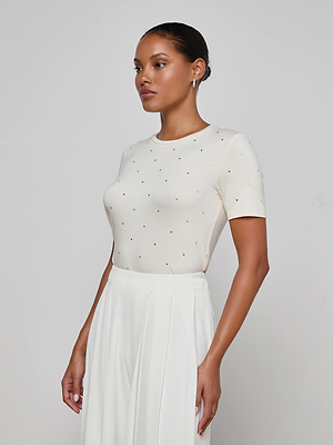 A woman with slicked-back hair poses in profile against a light background, wearing the Stormy Embellished Tee—a fitted, cropped cream top with small black dots—paired with high-waisted white pleated pants.