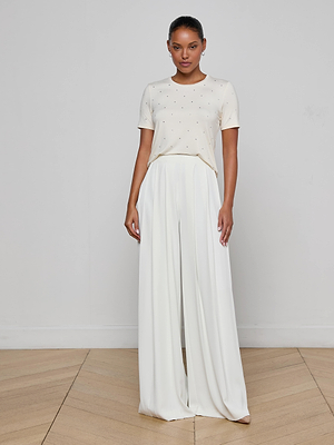 A woman stands indoors on a wooden floor, facing forward against a plain white wall. She wears the Stormy Embellished Tee paired with high-waisted, wide-leg white pants.