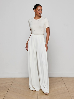 A woman stands against a white wall wearing the Stormy Embellished Tee, which features subtle crystal sparkle, paired with high-waisted, wide-leg vintage white trousers. Her slicked-back hair and neutral-toned heels complete the look.