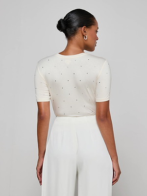 A person with hair in a neat bun, shown from behind against a light gray background, wears the Stormy Embellished Tee—a cream cropped short-sleeve top with small dots—paired with white high-waisted pants.