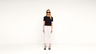 A woman wearing black sunglasses, a black t-shirt, white pants, and black heels stands against a plain white background, facing forward.