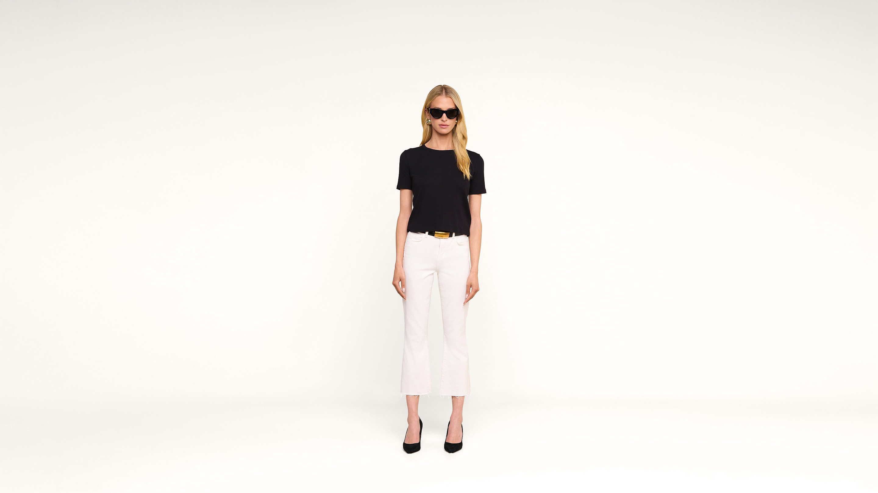 A woman wearing black sunglasses, a black t-shirt, white pants, and black heels stands against a plain white background, facing forward.