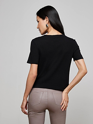 A person with long blonde hair, wearing the Stormy Cropped Cotton Tee in black—a semi-relaxed fit organic cotton top—and white pants, stands facing away from the camera against a plain, light background.