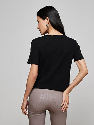 A woman with long dark hair is shown from the back, wearing the Stormy Cropped Cotton Tee in black, taupe pants, and silver hoop earrings, standing against a plain light gray background.