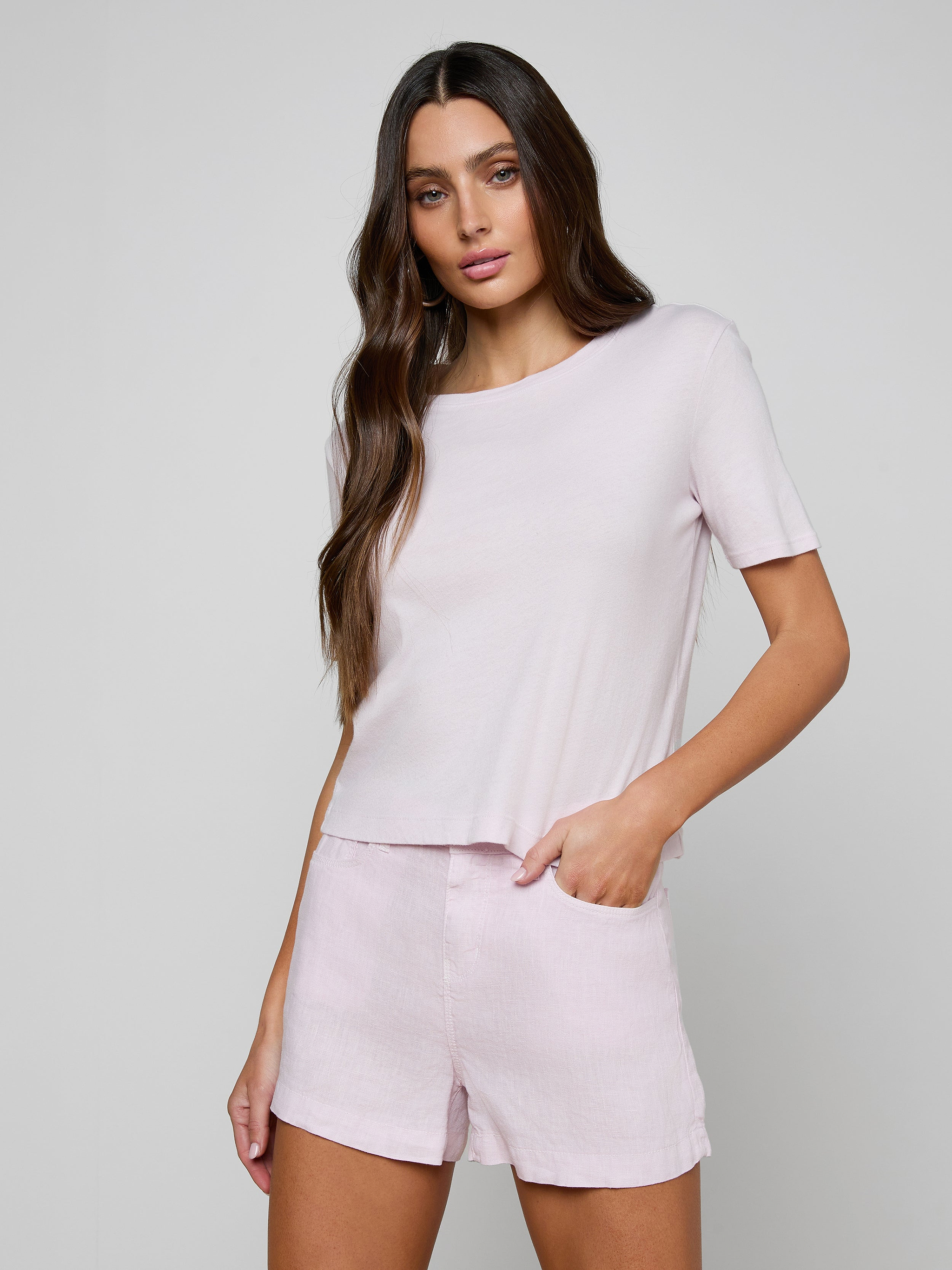 A woman with long brown hair wears the Stormy Cropped Cotton Tee in light pink—a semi-relaxed fit, cropped t-shirt—paired with matching shorts, standing with one hand in her pocket against a plain background.
