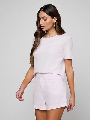 A woman with long brown hair models the Stormy Cropped Cotton Tee in pale pink, featuring organic cotton and a semi-relaxed fit, paired with matching high-waisted shorts, standing against a light gray background, looking down and to the side.