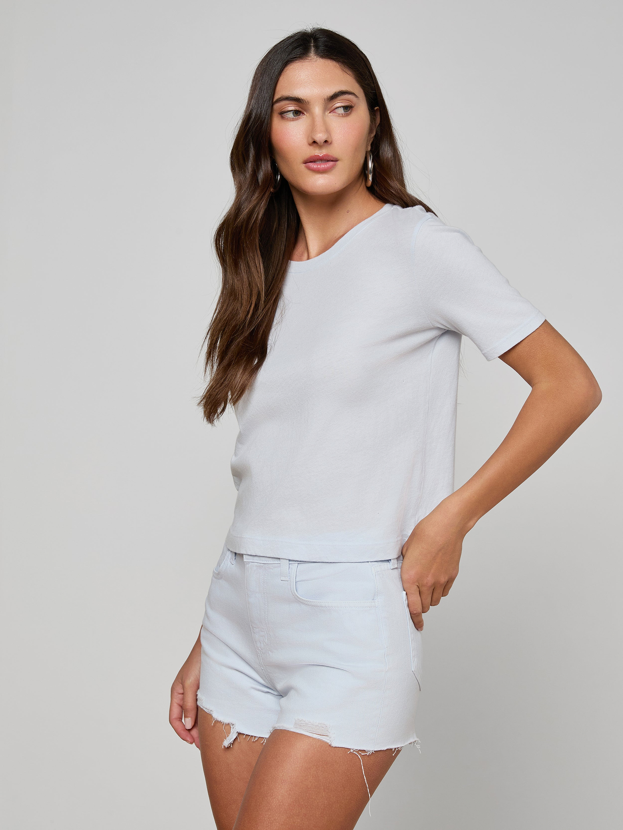 A woman with long brown hair wears the Stormy Cropped Cotton Tee, a plain white organic cotton shirt in a semi-relaxed fit, paired with light denim shorts. She stands against a plain light background, gazing slightly to the side with one hand near her shorts pocket.