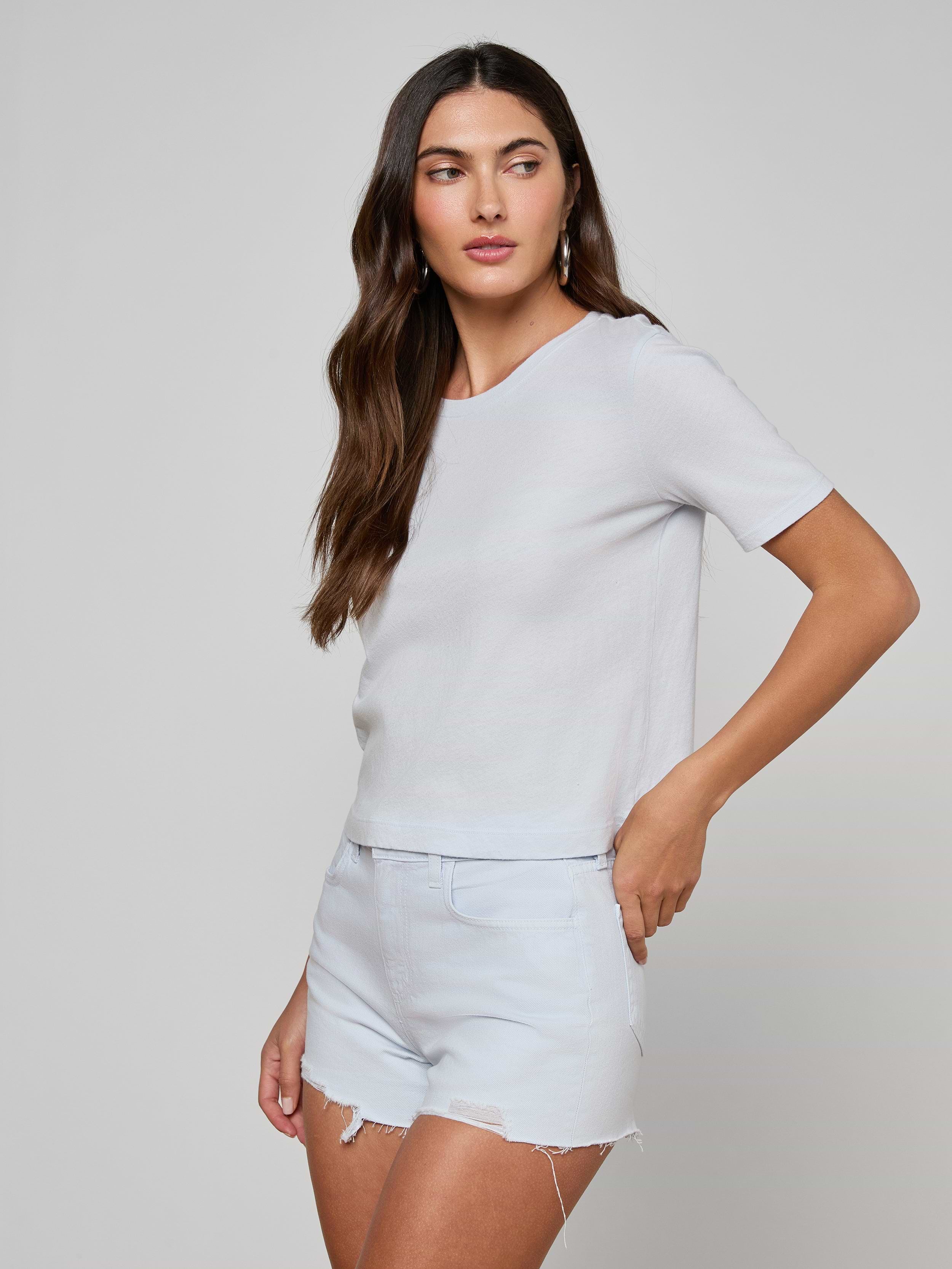 A woman with long brown hair wears the Stormy Cropped Cotton Tee, a plain white organic cotton shirt in a semi-relaxed fit, paired with light denim shorts. She stands against a plain light background, gazing slightly to the side with one hand near her shorts pocket.