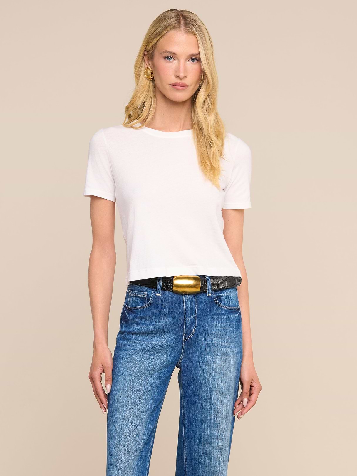 A woman stands against a plain beige background, wearing the Stormy Cropped Cotton Tee in white—featuring a semi-relaxed fit and crafted from organic cotton—paired with blue jeans, a black belt with a gold buckle, and gold hoop earrings. She has blonde wavy hair and a neutral expression.