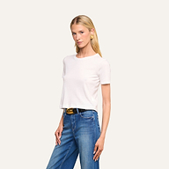 Una mujer con el pelo largo y rubio, que lleva la camiseta corta de algodón «Stormy» en color blanco —confeccionada en algodón orgánico y con un corte semiholgado—, combinada con unos vaqueros azules, un cinturón negro con hebilla dorada y pendientes dorados, posa frente a un fondo beige.